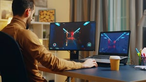 A man sitting at a desk gaming late at night on a dual-screen setup with a desktop monitor and a laptop, highlighting excessive screen time habits