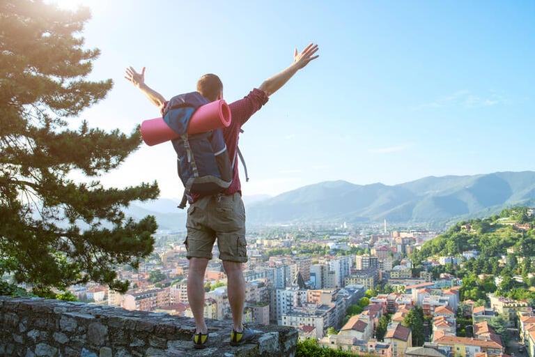 A traveler with a backpack stands on a scenic overlook above a city surrounded by mountains, arms raised in celebration