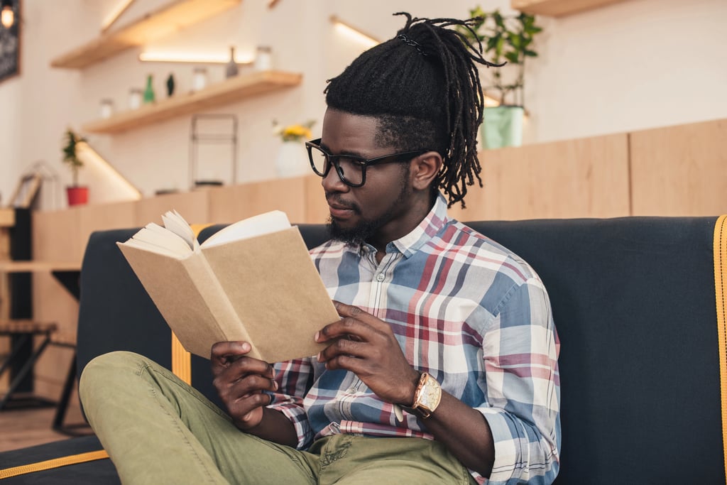 A Black man reading a book while sitting on a sofa in a cafe.