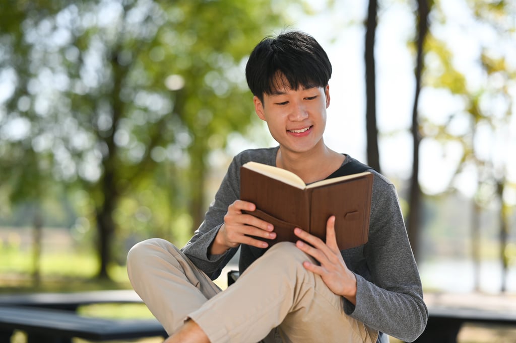 A happy young Asian man reading an interesting book while sitting bench in sunny summer park.