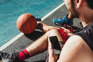 Two basketball players sitting on an outdoor court, holding their smartphones during a break with a basketball resting nearby
