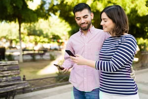 A smiling couple standing in a park, looking at a smartphone together, representing how online dating apps have transformed the way people meet and connect