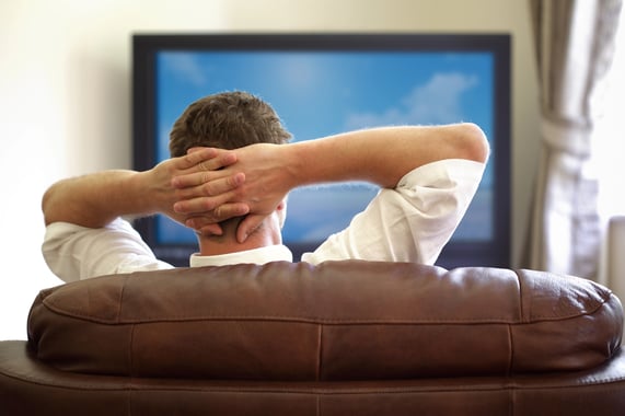 Man sitting on a sofa watching one of the best tv shows of the 21st century with hands folded behind his head.