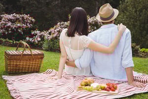 A couple sitting on a picnic blanket in a scenic garden, sharing a relaxed outdoor date with a basket and fresh food spread nearby