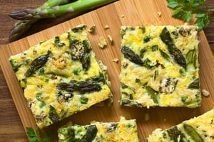 Asparagus and goat cheese frittata slices on a wooden board with a hand holding a playing card above.