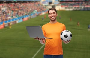 A smiling man in an orange shirt holding a laptop and a soccer ball, standing in front of a blurred stadium background to represent streaming sports.