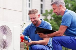 Two HVAC technicians in blue uniforms are diagnosing an outdoor heat pump unit using a manifold gauge set and a clipboard.