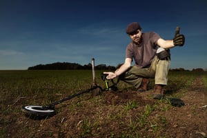 Man sitting in an open field using a metal detector, exploring the ground for buried coins and artifacts in a rural outdoor setting.