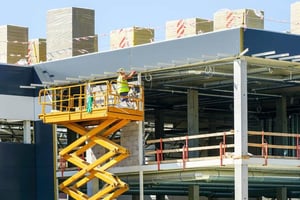A construction worker in a high-visibility vest and hard hat operates a yellow scissor lift to install grey exterior cladding on a large industrial building under construction.