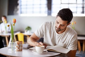 Man sitting at a table writing in a notebook beside a cup of coffee and flowers, representing simple weekly habits for staying organized and making time for reflection.