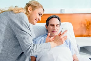 A woman smiling as she shows a smartphone to a man lying in a hospital bed, illustrating a successful way to get care for a loved one.