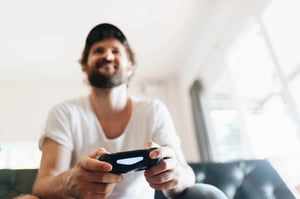 A smiling, bearded man holding a game controller while sitting on a couch, capturing why the best game days still feel simple.